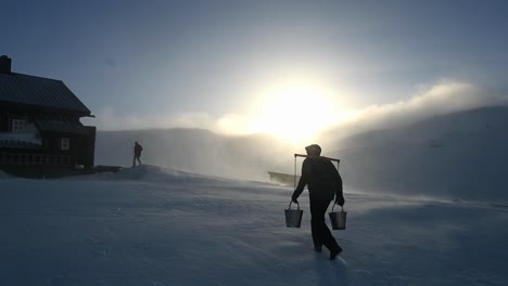 Silueta-De-Un-Hombre-Que-Lleva-Dos-Cubos-De-Agua-En-El-Hombro-De-Una-Montaña-Nevada-Hacia-La-Cabaña