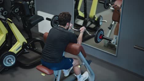 Un-Hombre-Motivado-Hace-Ejercicios-De-Curl-Con-Barra-De-Bíceps-Sentado-Frente-Al-Espejo