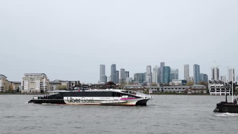 Uber-Boat-Reversing-From-Greenwich-Pier-With-Canary-Wharf,-Isle-Of-Dogs-Cityscape-In-Background