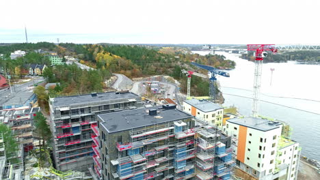 Vista-Aérea-De-Un-Sitio-De-Construcción-Con-Una-Gran-Grúa-Roja-Brillante-En-Funcionamiento-Cerca-Del-Río