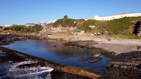 Calm-marine-tide-pool-with-people-swimming-at-sunrise,-Hermanus