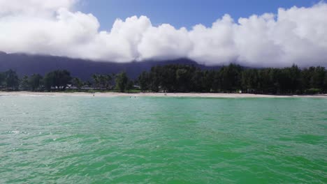 Imágenes-De-Drones-Recorriendo-Una-Playa-De-Arena-Blanca-Con-Agua-Turquesa-En-Primer-Plano-Y-Exuberante-Vegetación,-Montañas-Moradas-Y-Cielo-Azul-Con-Nubes-Blancas-En-El-Fondo-Oahu-Hawaii