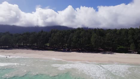 ascending-drone-footage-starting-with-the-turquoise-water-of-Waimanalo-Bay-to-white-sand-beach,-lush-forest,-purple-mountains-and-blue-sky-with-puffy-white-clouds