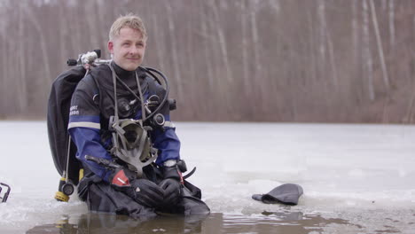 Man-in-dry-suit-and-technical-dive-gear-sits-on-edge-ice-lake-hole-chatting