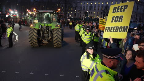 A-protestor-holds-up-a-placard-that-reads,-“Beep-for-freedom”-as-a-convoy-of-tractors-arrive-on-Parliament-Square-at-dusk-and-a-line-of-police-officers-keep-people-off-the-road