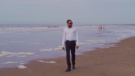 A-male-man-black-latino-model-in-a-suit-walks-on-the-beautifil-beach-seashore-with-sunglasses-in-the-Netherlands,-the-Hague