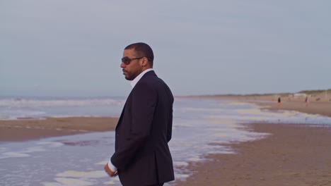 A-male-man-black-latino-model-in-a-suit-walks-on-the-picturesque-beach-and-takes-off-his-jacket-at-the-seashore-with-sunglasses-in-the-Netherlands,-the-Hague