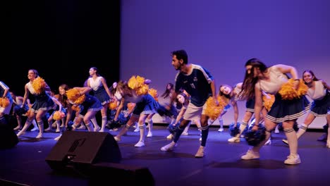 Grupo-De-Porristas-Con-Dos-Chicos-Haciendo-Una-Coreografía-En-Un-Escenario-Durante-Una-Velada-De-Gala,-Vestidos-De-Azul-Y-Blanco-Con-Pompones-Amarillos-Y-Azules.