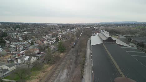 Aerial-drone-view-of-train-commuting-with-neighborhood-on-one-side-of-tracks-and-manufacturing-plant-on-the-other