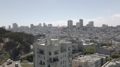 Drone-circles-flag-topped-building-in-San-Francisco,-revealing-Coit-Tower-and-downtown-skyline