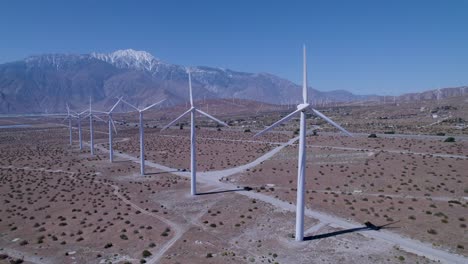Slow-tracking-drone-shot-showcases-windmills-in-the-desert-with-a-snow-capped-mountain-in-the-background,-along-with-more-windmills-in-the-distance
