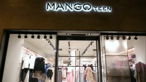 Pedestrians-walk-past-the-Spanish-multinational-clothing-brand-Mango,-Mango-Teen-store,-during-nighttime-in-Spain