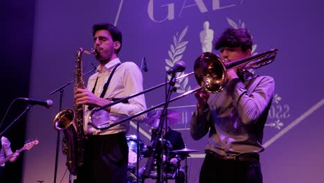 young-saxophonist-and-trumpet-player-at-a-gala-concert,-soft-lighting,-drums-behind-and-bass-player,-singers-on-the-left