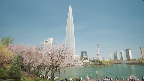 Lotte-World-Tower-and-People-Walking-on-Walkways-by-Seokchon-Lake-of-Songpa-Naru-Park-In-Seoul,-South-Korea---high-angle-wide-shot