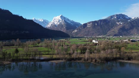 Clip-De-Drone-Que-Muestra-Majestuosas-Montañas-Cubiertas-De-Nieve-Con-Exuberante-Tierra-Verde-Y-Un-Lago-En-Primer-Plano