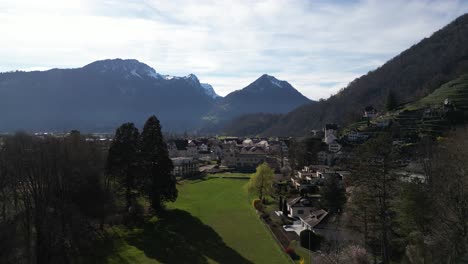 Drone-clip-showing-Swiss-village-with-white-buildings,-surrounding-lush-grassland-and-distant-snowy-mountains