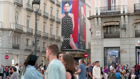 Crowds-fill-Puerta-del-Sol-with-the-backdrop-of-a-fashion-retail-billboard-from-El-Corte-Inglés,-Madrid,-Spain