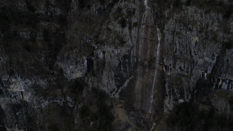 Rising-drone-clip-of-high-mountain-waterfall-showing-steep-rock-face-and-water's-edge-at-base-of-cliff