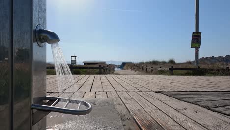 Agua-Potable-Que-Fluye-De-Una-Brillante-Fuente-De-Acero-Inoxidable-Que-Cae-Sobre-La-Madera-De-Acceso-Al-Mar-De-Un-Pueblo-Con-Barandillas-De-Madera-Antiguas,-Primer-Plano-Bloqueado,-Pontevedra,-Galicia,-España