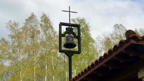 Campana-De-Latón-Se-Balancea-Ligeramente-En-Un-Soporte-Rectangular-Debajo-De-Una-Delgada-Cruz-En-Un-Día-Ventoso-En-Las-Montañas