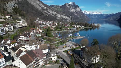 Drone-clip-showing-white-buildings-in-small-lakeside-village-in-Switzerland,-with-snowy-mountains-in-the-background