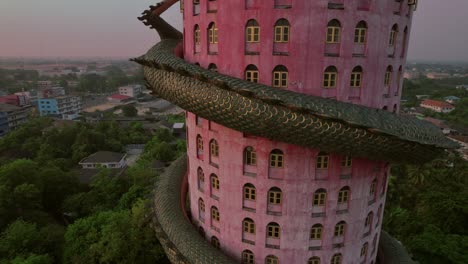 Rotating-aerial-view-of-the-"Dragon-Temple"-Wat-Sam-Phran,-an-extraordinary-17-story-cylindrical-pink-temple-embellished-with-a-massive-green-dragon-wrapping-around-the-edifice