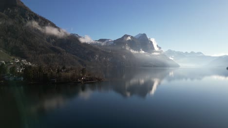 Drohnenclip,-Der-Das-Seeufer-Mit-Nebligen-Und-Schneebedeckten-Bergen-An-Einem-Strahlend-Sonnigen-Tag-Zeigt