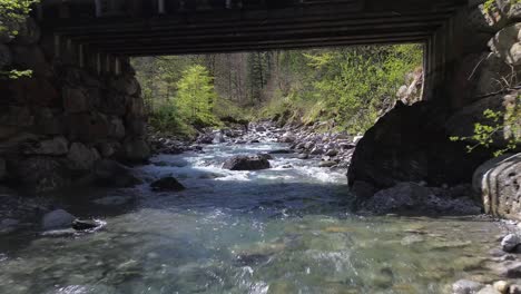 Drone-Fly-above-River-underneath-Bridge-in-Idyllic-Mountain-Valley-in-Austria,-Europe