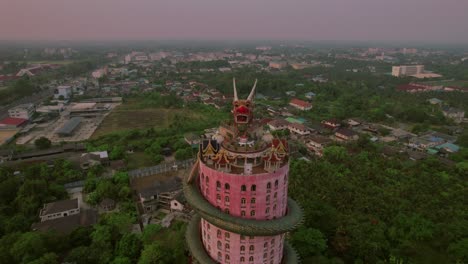 Aerial-rotation-showcasing-the-"Dragon-Temple"-Wat-Sam-Phran,-an-impressive-17-story-cylindrical-pink-temple-embellished-with-a-massive-green-dragon-encircling-its-structure,-situated-in-Bangkok