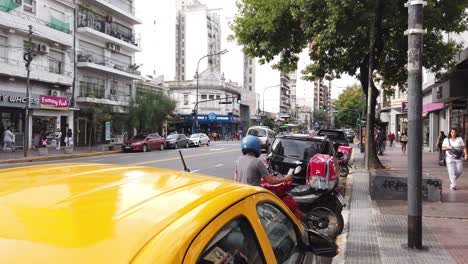 Delivery-motorbike-driver-waits-outside-commercial-area-in-traffic-latin-busy-avenue-Pedidosya,-yellow-black-taxi-of-buenos-aires-city-metropolitan-area