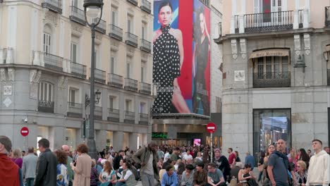 La-Gente-Se-Reúne-En-La-Puerta-Del-Sol,-Enmarcada-Por-El-Reconocible-Cartel-De-Venta-De-Moda-Al-Fondo,-En-Madrid,-España.