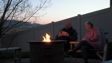 Senior-couple-at-a-fire-pit,-wife-demands-attention-while-husband-is-on-a-smartphone-device