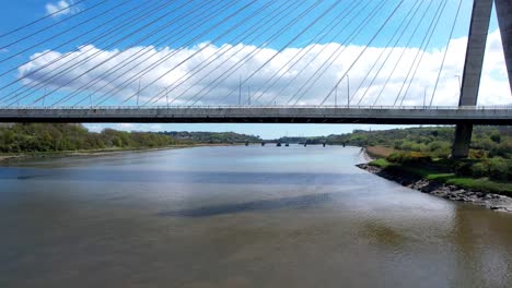 Drone-Pasando-Bajo-El-Puente-Colgante-Sobre-El-Río-Suir-En-Waterford-Irlanda-Volando-Bajo-Sobre-El-Río-Y-El-Antiguo-Puente-Ferroviario-Apareciendo-A-La-Vista