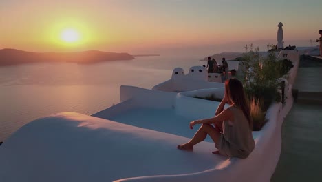 Una-Mujer-Se-Sienta-Tranquilamente-En-La-Pared-De-Una-Casa-En-La-Isla-De-Santorini,-Contemplando-La-Colorida-Puesta-De-Sol-Sobre-El-Mar.