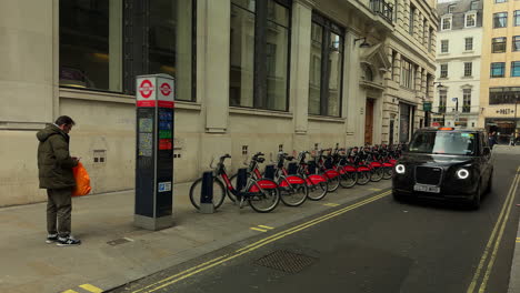 Santander-Hire-BIke-station,-Taxi-drives-past,-pedestrian,-Central-London,-Wide-shot,-Day