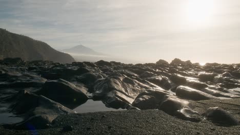 Etérea-Playa-De-Tenerife-Al-Amanecer-Con-Arena-Negra-Y-Roca-Volcánica-Y-El-Brumoso-Pico-Del-Teide-Al-Fondo,-Islas-Canarias