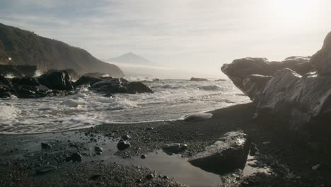 Ethereal-black-sand-beach-and-Pico-del-Teide-mountain-at-distance,-misty-shore-with-smooth-waves-at-dawn,-Tenerife-Canary-Islands