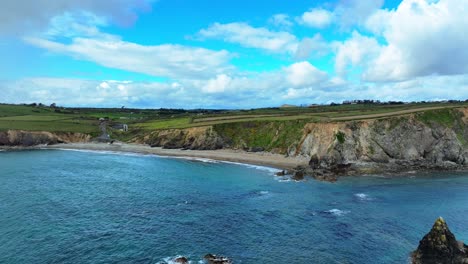 Drone-flying-over-rocks-and-crystal-clear-water-along-beach-with-people-walking-to-sea-cliffs-in-Waterford-Ireland-on-a-spring-day