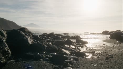 ätherischer-Schwarzer-Sandstrand-Mit-Pico-Del-Teide-In-Der-Ferne,-Neblige-Felsen-Mit-Sanften-Wellen-Im-Morgengrauen,-Teneriffa,-Kanarische-Inseln