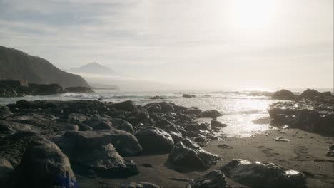 Etérea-Playa-De-Arena-Negra-Y-Pico-Del-Teide-A-Distancia,-Rocas-Brumosas-Con-Suaves-Olas-Al-Amanecer,-Tenerife-Islas-Canarias