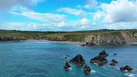 Drone-static-shot-jagged-dark-rocks-on-a-deep-blue-sea-in-a-bay-on-the-Copper-Coast-Waterford-Ireland-establishing-shot-of-stunning-coastline