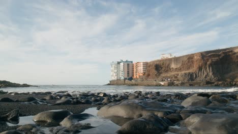 Black-sand-pebbles-beach-shoreline-with-cliff-and-tall-buildings-under-tranquil-blue-sky,-Playa-de-la-Arena,-Tenerife,-low-angle