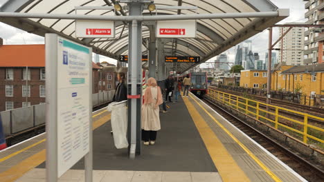 Synchronisierte-Ankunft-Von-DLR-Zügen-An-Einer-Oberirdischen-Station-Im-Osten-Londons,-Während-Pendler-Vor-Der-Kulisse-Der-Skyline-Des-Londoner-Finanzviertels-Warten