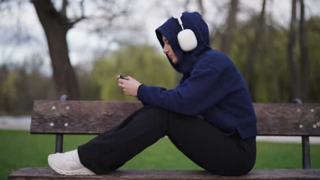 Alone-woman-sit-on-wooden-park-bench-and-use-smartphone-with-wireless-headset