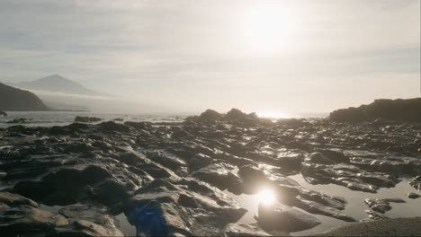 Sonnenreflexion-Auf-Wasserpfützen-Und-Schwarzem-Vulkangestein,-Strand-Auf-Teneriffa-Und-Pico-Del-Teide-Unter-Klarem-Himmel,-ätherisch,-Kanarische-Inseln,-Langsame-Schwenk