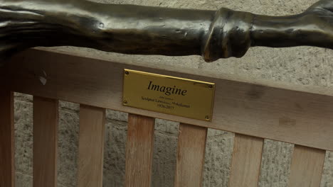 Statue-of-John-Lennon-on-a-bench,-Imagine,-Carnaby-Street,-London,-Day,-People-walk-past,-Close-up-on-plaque