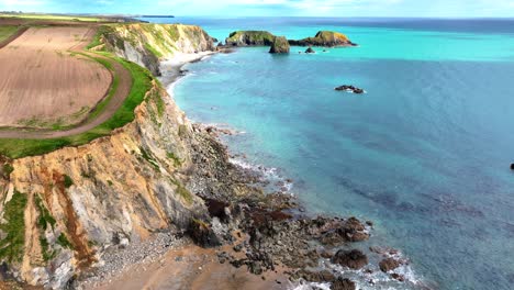 Drones-Volando-Sobre-Promontorios-Con-Acantilados-Arenosos-Erosionando-El-Mar-Y-Desprendimientos-De-Rocas-En-La-Playa-Con-Aguas-Cristalinas-Y-Colores-Azules-Profundos-Costa-De-Cobre-En-Waterford,-Irlanda,-En-Una-Tarde-De-Primavera