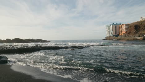 Hotel-on-cliff-under-serene-sky-in-Tenerife,-ocean-waves-on-rocky-black-sand-beach,-Canary-Island