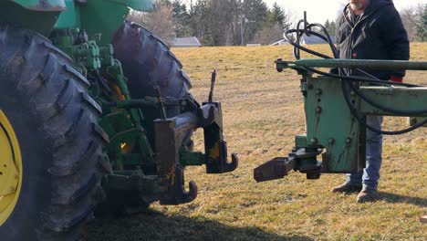 Agricultor-Sujetando-Un-Arado-Al-Enganche-De-Tres-Puntos-En-La-Parte-Trasera-Del-Tractor,-Las-Mangueras-Hidráulicas-Son-Visibles-Pero-No-Están-Conectadas