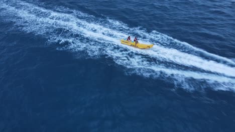 Pareja-Montando-Un-Trineo-Banana-En-Agua-De-Mar-Arrastrada-Por-Una-Moto-Acuática,-Drone-Sigue-El-Disparo